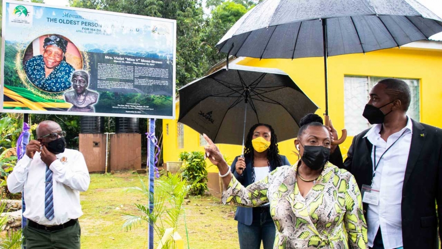 The Minister of Culture, Gender, Entertainment and Sport, the Honourable Olivia Grange, points to the storyboard erected at the home of the late supercentenarian, Mrs Violet Moss-Brown, in Duanvale, Trelwany.  Mrs Moss-Brown, who lived for 117 years and 189 days, was the record holder for world’s ‘Oldest living person’. Also pictured are the Mayor of Falmouth, His Worship Councillor Colin Gager (left) and Member of Parliament for Northern Trelawny, Tova Hamilton (partially hidden) and Constable Renaldo Riley.