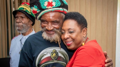 The Minister of Culture, Gender, Entertainment and Sport, the Honourable Olivia Grange, embraces  Edward Fray, a survivor of the 1963 Coral Gardens incident after the formal establishment of the Trust for victims which will be administered by the Administrator General of Jamaica.  Also pictured is Isaac Wright, another survivor.